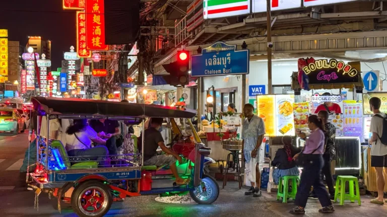 A busy Bangkok street at night with a tuk-tuk, neon signs, food stalls and pedestrians.