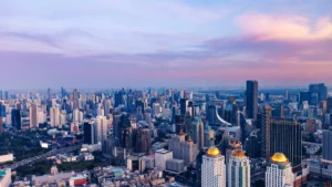 A dense city skyline stretches beneath a pink and blue sunset, with high-rise towers in the foreground.