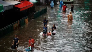 Thailand’s national catastrophe insurance scheme makes first payout People wade through floodwater carrying bags and belongings beside a market.
