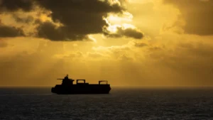 Silhouette of a cargo ship on the sea beneath a golden, cloud-filled sunset.